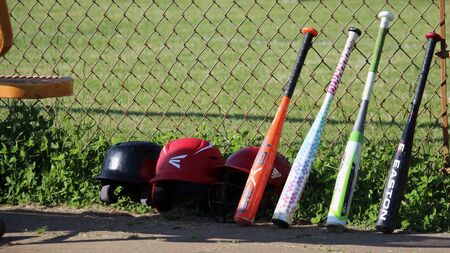 Row of kids’ baseball helmets and baseball bats leaning against a fence