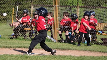 Young baseball player swings a bat at a baseball over home plate. A boy mimics by swinging his own bat, while other teammates watch from a bench in the dugout.