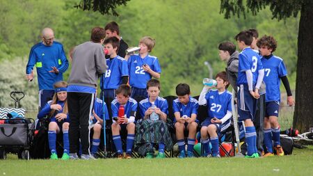 Youth soccer team resting on bench during half time of game, listening to coaches and drinking water.